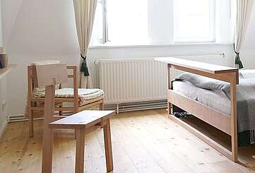 Moderne Schlafzimmermöbel aus hellem Holz stehen in einem lichtdurchfluteten Zimmer. Cremefarbene Vorhänge und der Holzfußboden tragen zur Natürlichkeit bei.