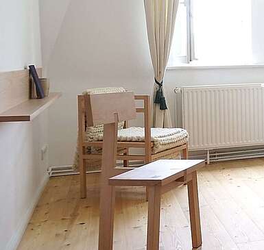 Moderne Schlafzimmermöbel aus hellem Holz stehen in einem lichtdurchfluteten Zimmer. Cremefarbene Vorhänge und der Holzfußboden tragen zur Natürlichkeit bei.