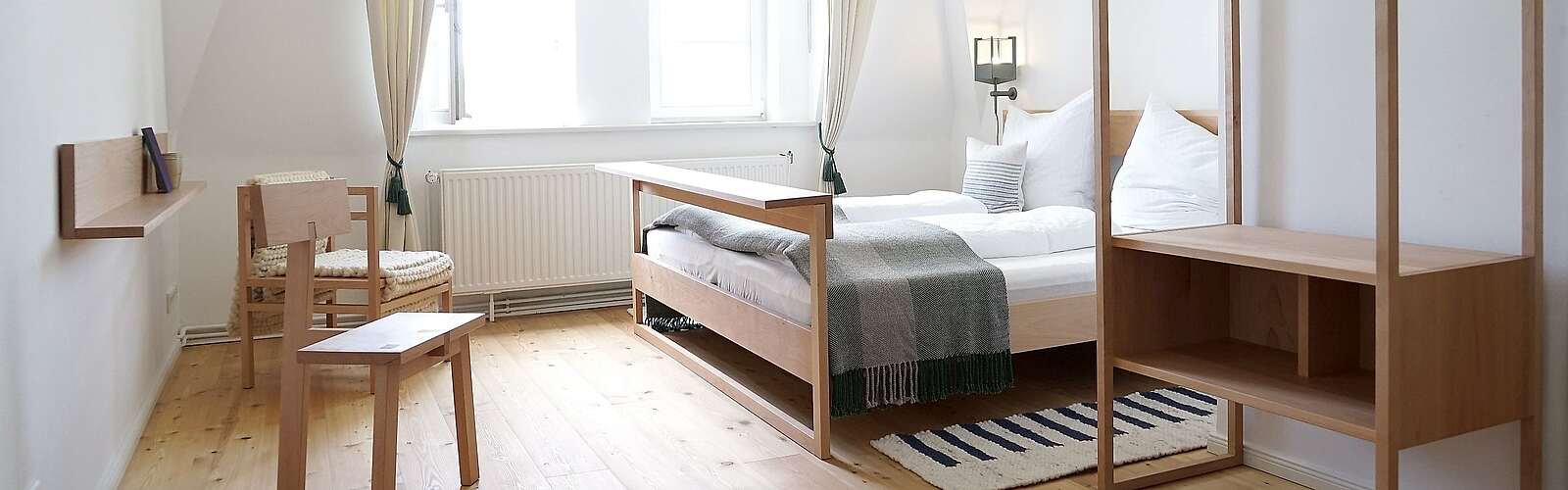 Moderne Schlafzimmermöbel aus hellem Holz stehen in einem lichtdurchfluteten Zimmer. Cremefarbene Vorhänge und der Holzfußboden tragen zur Natürlichkeit bei.