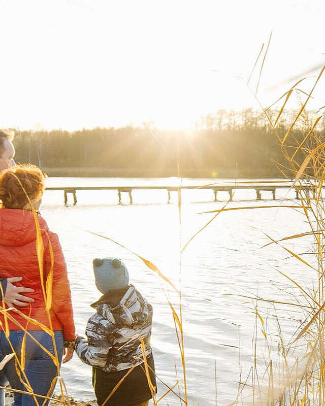 Eine Familie aus Vater, Mutter und Kind steht in der Abenddämmerung am Seeufer.