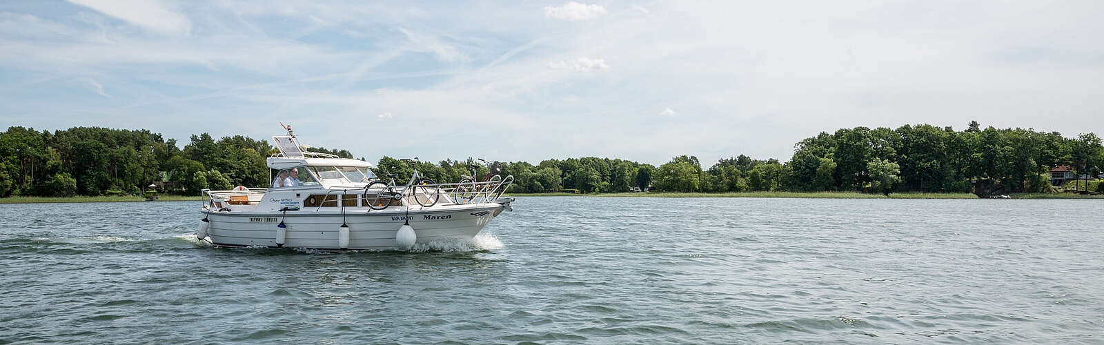 Ein sportliches Hausboot gleitet über den See. Am Ufer sind grüne Bäume zu erahnen, der Himmel strahlt blau.