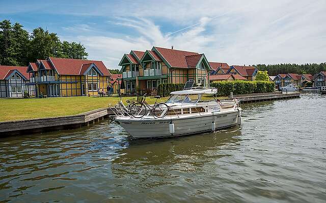 Ein Hausboot fährt auf dem Kanal vor den Ferienhäusern des Hafendorfs Rheinsberg entlang. Zwei Menschen sind an Bord.