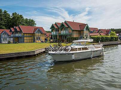 Ein Hausboot fährt auf dem Kanal vor den Ferienhäusern des Hafendorfs Rheinsberg entlang. Zwei Menschen sind an Bord.