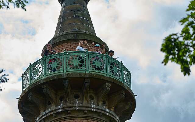 Das von unten aufgenommene Foto zeigt oben auf der Galerie der Siegessäule stehende Frauen, die lachend nach unten winken.