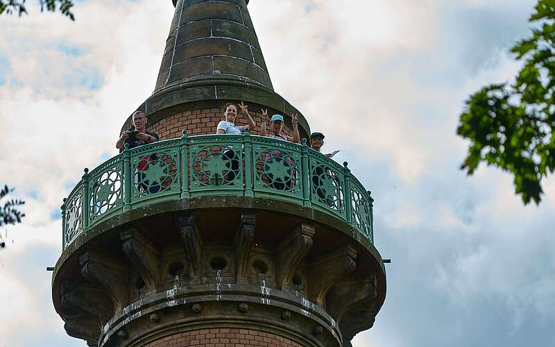Das von unten aufgenommene Foto zeigt oben auf der Galerie der Siegessäule stehende Frauen, die lachend nach unten winken.