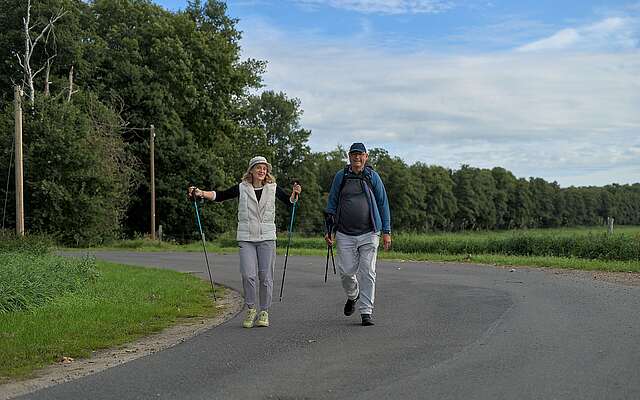 Ein älteres Ehepaar wandert in sportlicher Kleidung und mit Wanderstöcken in der Hand über eine asphaltierte Straße, die von grünen Wiesen und Bäumen gesäumt ist.