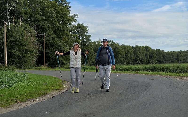 Ein älteres Ehepaar wandert in sportlicher Kleidung und mit Wanderstöcken in der Hand über eine asphaltierte Straße, die von grünen Wiesen und Bäumen gesäumt ist.