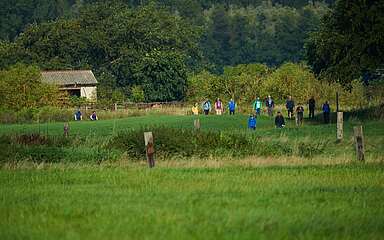 Aus der Ferne sieht man Menschen durch eine grüne Feldlandschaft wandern.