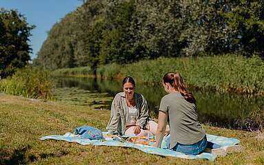 Zwei junge Frauen picknicken bei Sonnenschein am Ufer eines Kanals.