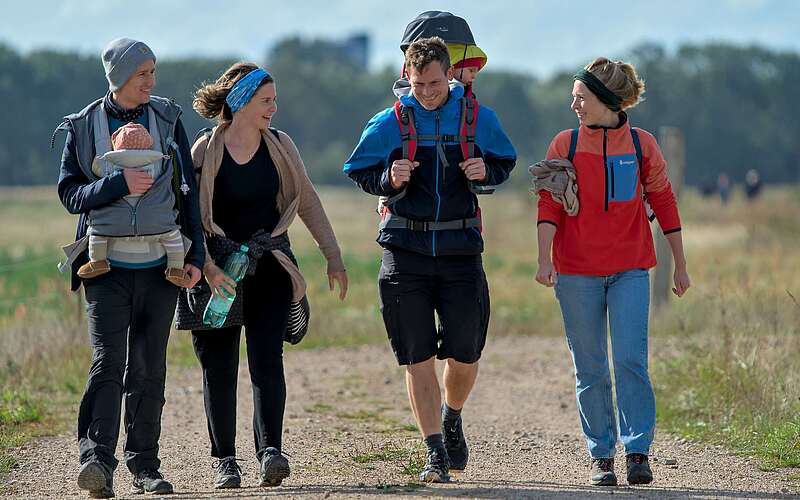 Zwei Familien mit Kleinkindern in Tragerucksäcken wandern über ein Feld.