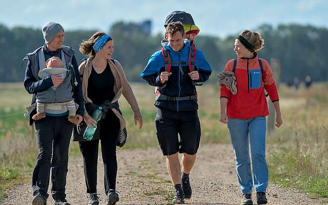 Zwei Familien mit Kleinkindern in Tragerucksäcken wandern über ein Feld.