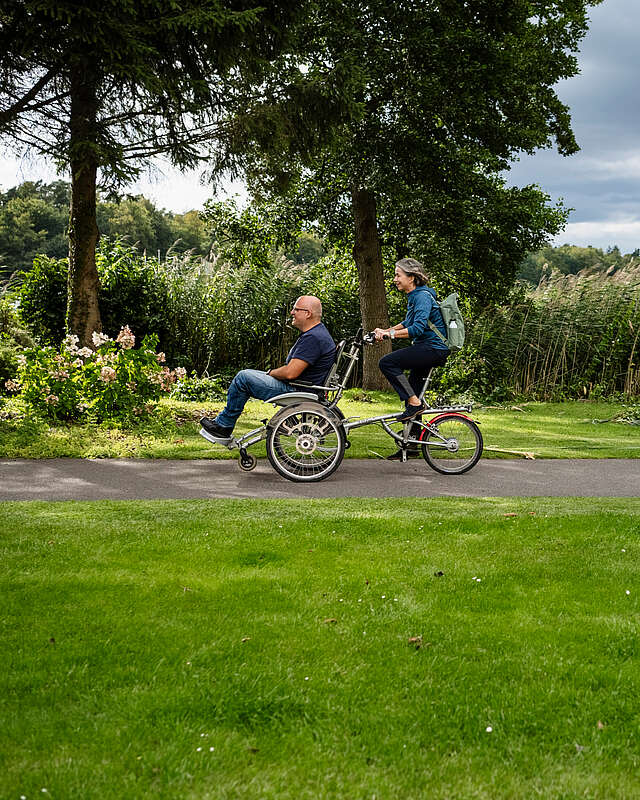 Ein Mann und eine Frau fahren gemeinsam auf dem Rollfiets über einen asphaltierten Weg, der auf der einen Seite von Uferschilf gesäumt wird. Die Frau tritt in die Pedale, der Mann hat auf dem vorderen Sitz Platz genommen.
