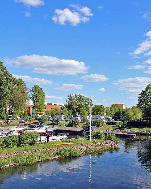 Boote fahren bei sommerlichem Wetter auf einem Kanal. Nach links führt eine kleine Wasserstraße zu Anlegeplätzen. Im Hintergrund sind Wohnmobile an Land zu erkennen.