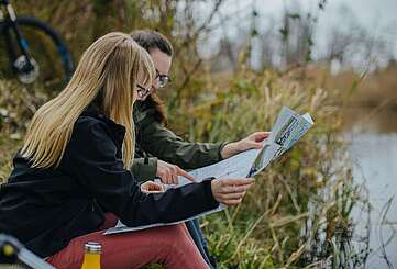 Zwei Frauen sitzen im Gras am Flussufer und schauen sich gemeinsam eine Radkarte an.