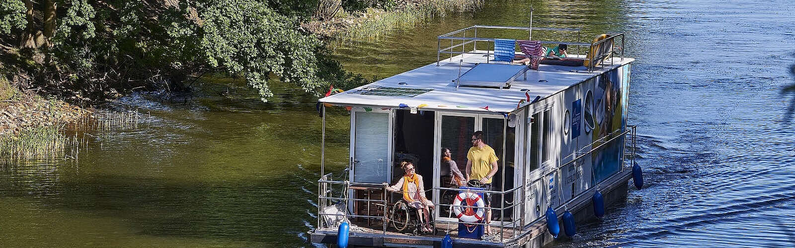 Ein Hausboot fährt in einen Kanal. Ein Mann steht hinter dem Steuer, eine Frau sitzt neben ihm im Rollstuhl. Oben auf dem Sonnendeck liegt ein Junge und liest.