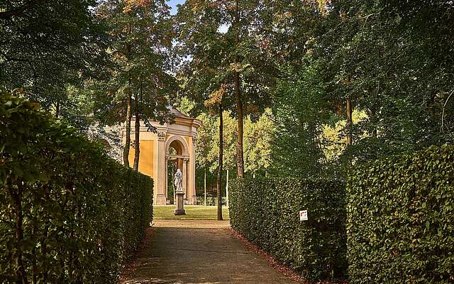 Weg zwischen hohen, geschnittenen Hecken mit einer Statue und einem kleinen Pavillon im Hintergrund