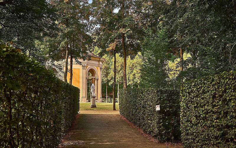 Weg zwischen hohen, geschnittenen Hecken mit einer Statue und einem kleinen Pavillon im Hintergrund