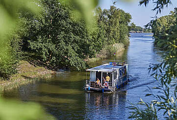Ein Hausboot fährt in einen Kanal. Ein Mann steht hinter dem Steuer, eine Frau sitzt neben ihm im Rollstuhl. Oben auf dem Sonnendeck liegt ein Junge und liest.