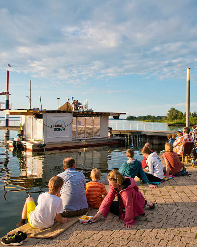 Zwei Musiker sitzen auf dem Dach des Traumschüffes, einer mobilen Bühne auf dem Wasser. Zuschauer sitzen am Ufer.