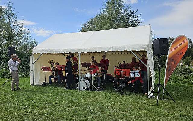 <p>Ein Mann fotografiert die unter einem Veranstaltungszelt sitzende, in rot gekleideten Mitglieder der Blaskapelle.</p>