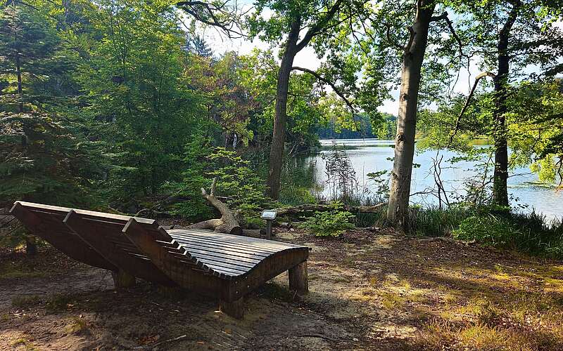 Holzliege auf einem Waldweg am Ufer eines Sees mit Bäumen und Wasser im Hintergrund