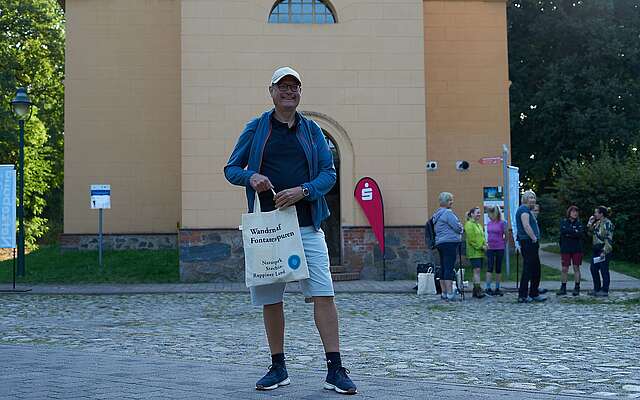 Ein Mann steht in sportlicher Kleidung vor der Temnitzkirche und hält einen Beutel mit Aufdruck des Naturparks Stechlin-Ruppiner Land in der Hand.