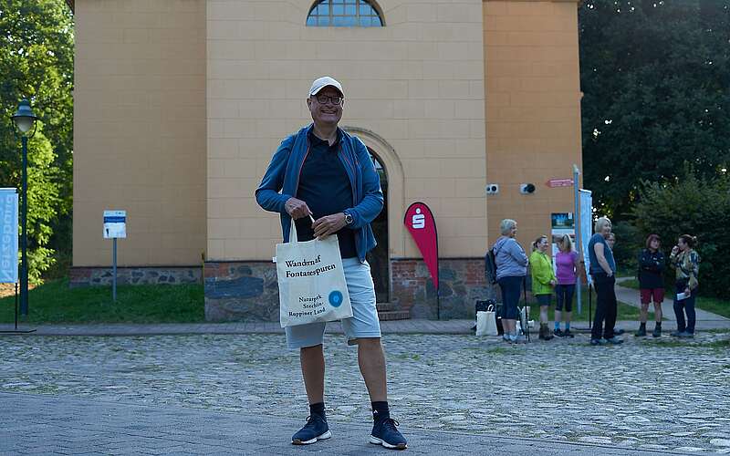 Ein Mann steht in sportlicher Kleidung vor der Temnitzkirche und hält einen Beutel mit Aufdruck des Naturparks Stechlin-Ruppiner Land in der Hand.