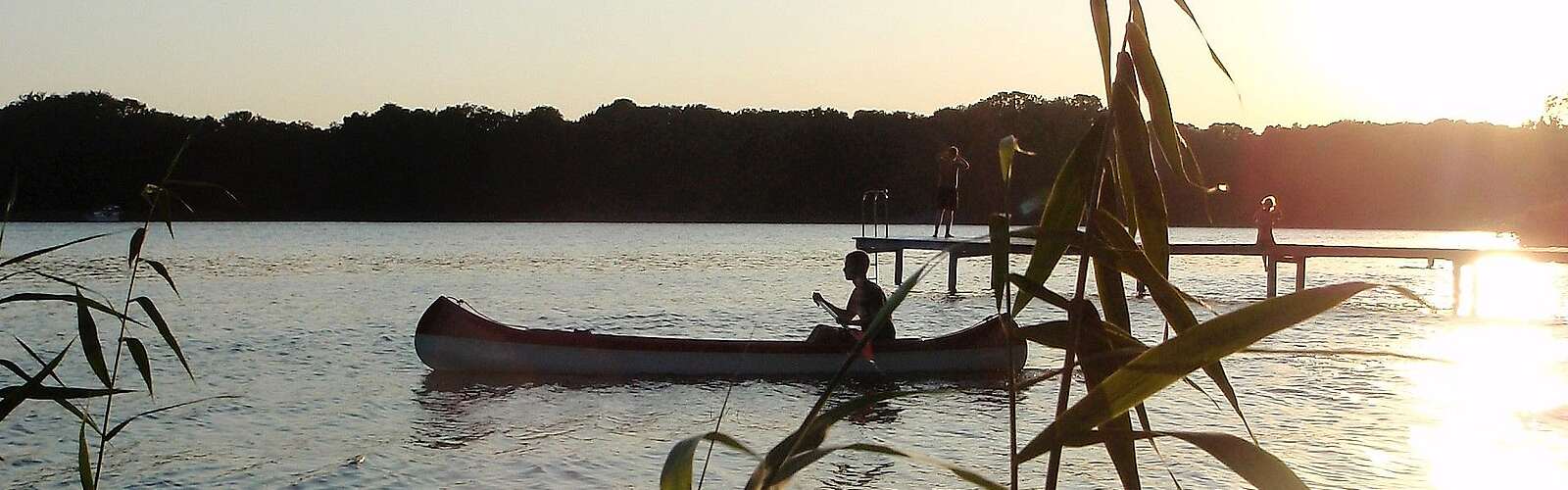 Eine Person fährt in der Abenddämmerung Kanu. Die untergehende Sonne spiegelt sich im Wasser.