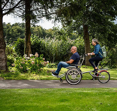 Ein Mann und eine Frau fahren gemeinsam auf dem Rollfiets über einen asphaltierten Weg, der auf der einen Seite von Uferschilf gesäumt wird. Die Frau tritt in die Pedale, der Mann hat auf dem vorderen Sitz Platz genommen.