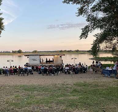 Schauspieler stehen auf dem Traumschüff, einer mobilen Bühne auf dem Wasser. Zuschauer schauen vom Ufer aus zu.