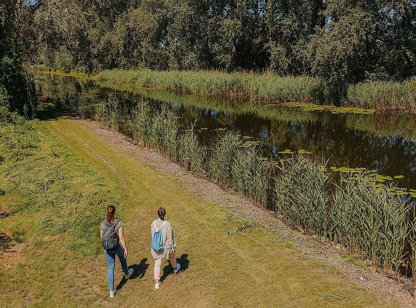 Zwei junge Frauen spazieren am grünen Ufer des Kremmener Kanals entlang.