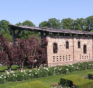 Ein Backsteingebäude steht am Rand von saftig grünen Rasenflächen, die von geschnittenen Hecken und bepflanzeten Beeten gesäumt sind. Im Hintergrund blühen Bäume.
