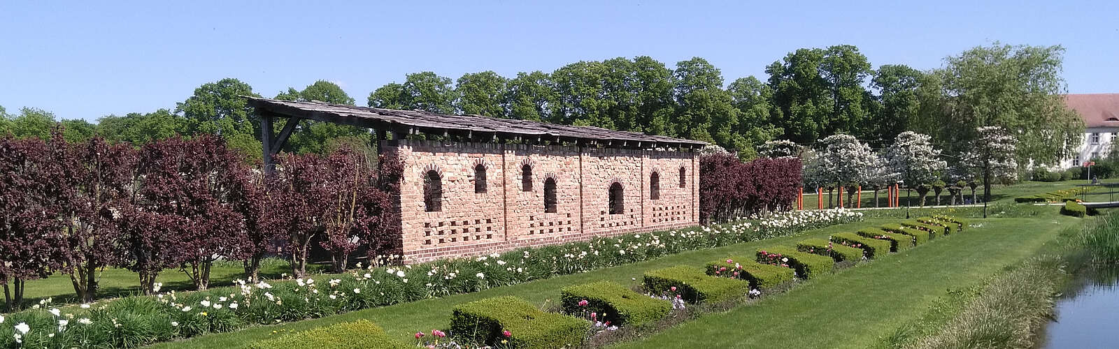 Ein Backsteingebäude steht am Rand von saftig grünen Rasenflächen, die von geschnittenen Hecken und bepflanzeten Beeten gesäumt sind. Im Hintergrund blühen Bäume.