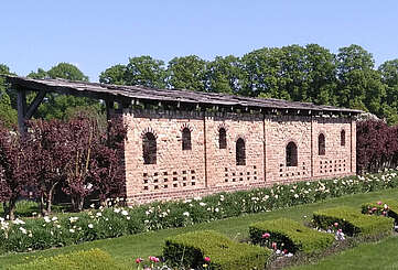 Ein Backsteingebäude steht am Rand von saftig grünen Rasenflächen, die von geschnittenen Hecken und bepflanzeten Beeten gesäumt sind. Im Hintergrund blühen Bäume.