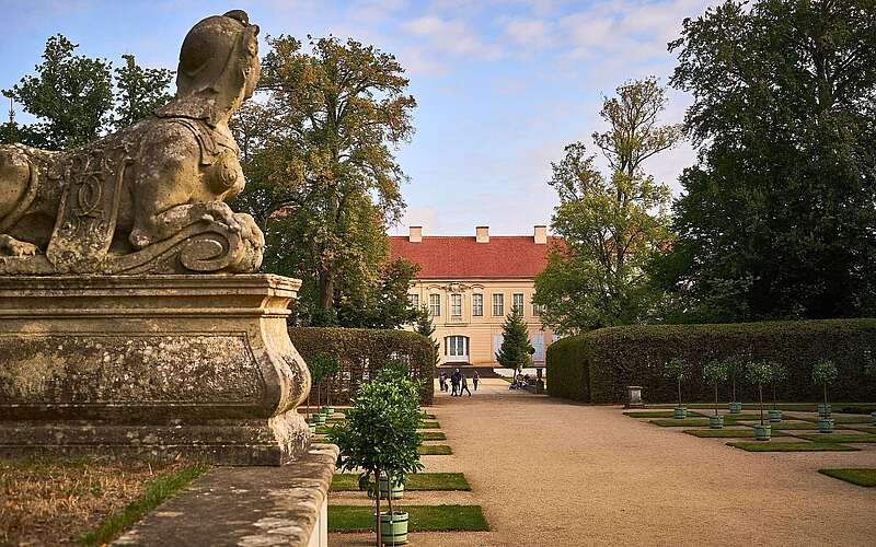 Steinerne Sphinx-Skulptur im Schlosspark mit Blick auf ein barockes Schloss und symmetrisch angelegte Gartenwege