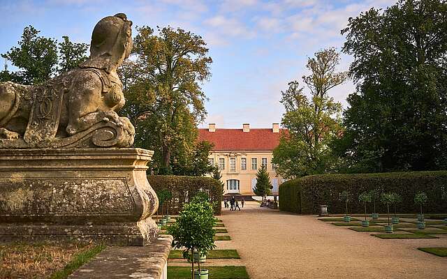 Steinerne Sphinx-Skulptur im Schlosspark mit Blick auf ein barockes Schloss und symmetrisch angelegte Gartenwege