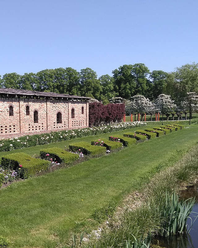 Ein Backsteingebäude steht am Rand von saftig grünen Rasenflächen, die von geschnittenen Hecken und bepflanzeten Beeten gesäumt sind. Im Hintergrund blühen Bäume.