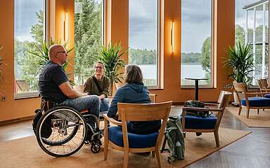 Ein Mann im Rollstuhl und zwei Frauen sitzen an einer Sitzgruppe im Foyer des Seehotel in Rheinsberg. Hinter ihnen kann man durch die großen Fenster den Grienericksee erkennen.