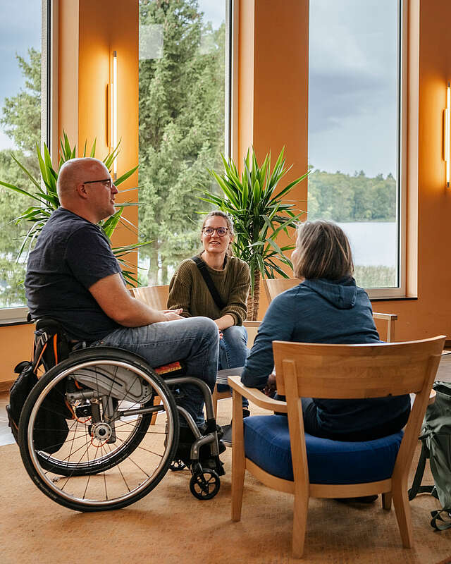 Ein Mann im Rollstuhl und zwei Frauen sitzen an einer Sitzgruppe im Foyer des Seehotel in Rheinsberg. Hinter ihnen kann man durch die großen Fenster den Grienericksee erkennen.