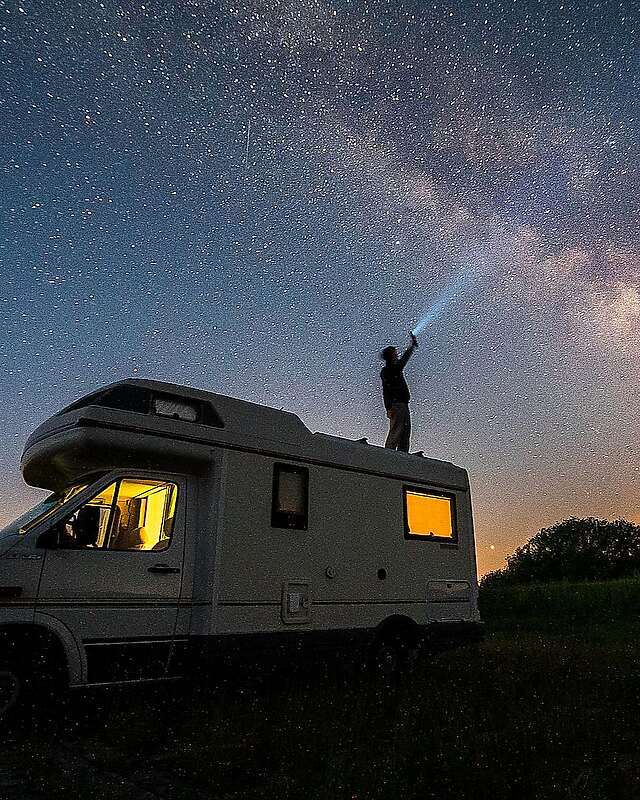 Ein Mann steht bei Nacht auf dem Dach seines Wohnmobils und hält eine Taschenlampe in Richtung Himmel. Der Lichtstrahl ist vor der ebenfalls deutlich sichtbaren Milchstraße zu erkennen.