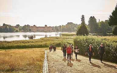 Gruppe von Menschen wandert auf einem Weg durch einen Park mit Schloss und See im Hintergrund