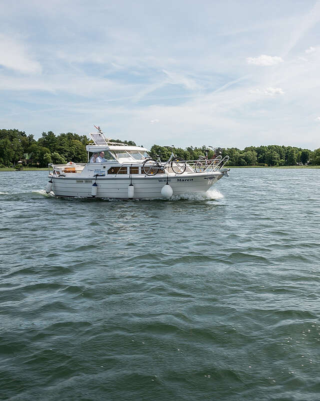 Ein sportliches Hausboot gleitet über den See. Am Ufer sind grüne Bäume zu erahnen, der Himmel strahlt blau.