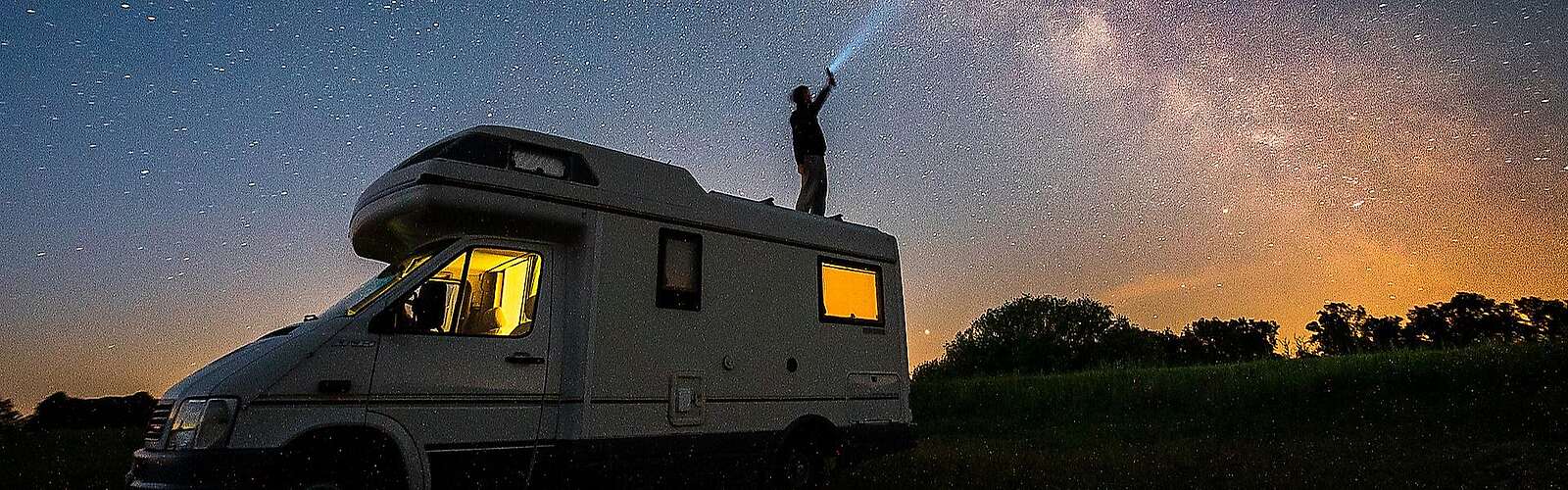 Ein Mann steht bei Nacht auf dem Dach seines Wohnmobils und hält eine Taschenlampe in Richtung Himmel. Der Lichtstrahl ist vor der ebenfalls deutlich sichtbaren Milchstraße zu erkennen.