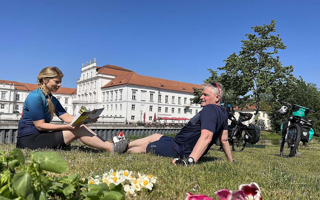 Eine Radfahrerin und ein Radfahrer sitzen in Sportkleidung auf einer Wiese. Sie hält eine Radkarte in der Hand und er schaut auf das hinter ihnen liegende Schloss Oranienburg. Neben ihnen stehen die Fahrräder mit Gepäcktaschen.