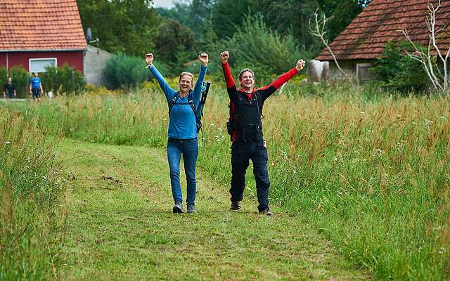 Eine Frau und ein Mann wandern über einen ins grüne Feld gemähten Weg. Sie strecken freudig die Arme in die Luft und lachen.