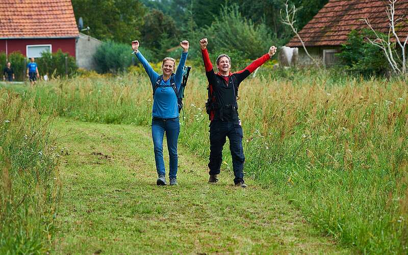 Eine Frau und ein Mann wandern über einen ins grüne Feld gemähten Weg. Sie strecken freudig die Arme in die Luft und lachen.