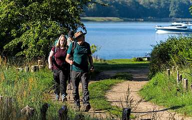 Mehrere Personen wandern bei sommerlichem Wetter einen Weg hinauf in Richtung Fotograf. Hinter ihnen sieht man einen See, auf dem ein Hausboot fährt.