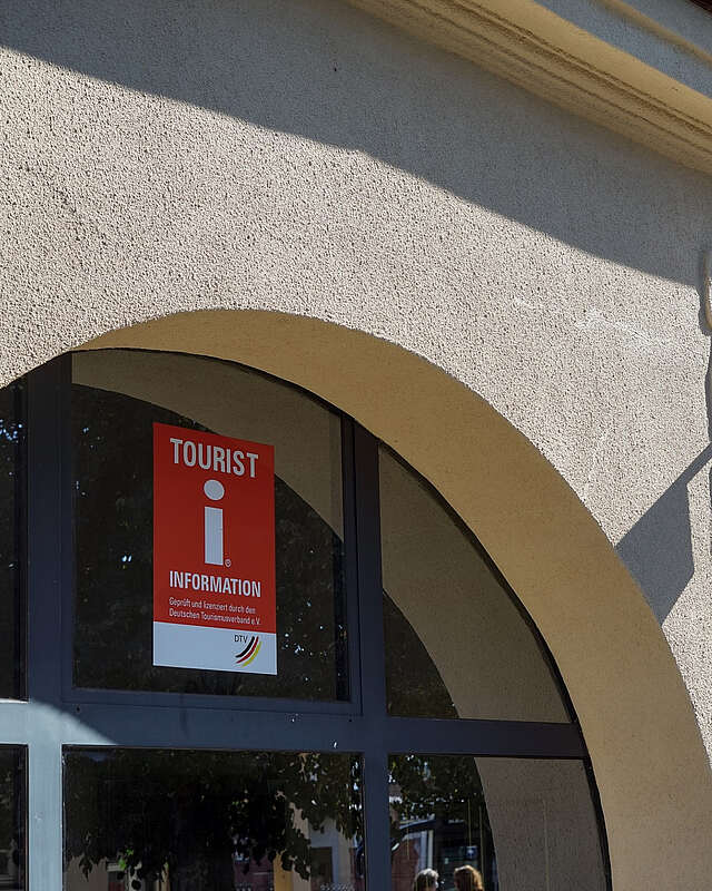 Eine Häuserfront mit oben abgerundeten Fenstern. An der Scheibe und an der verputzten Wand hängen Schilder mit dem roten "i" und eine Lampe.
