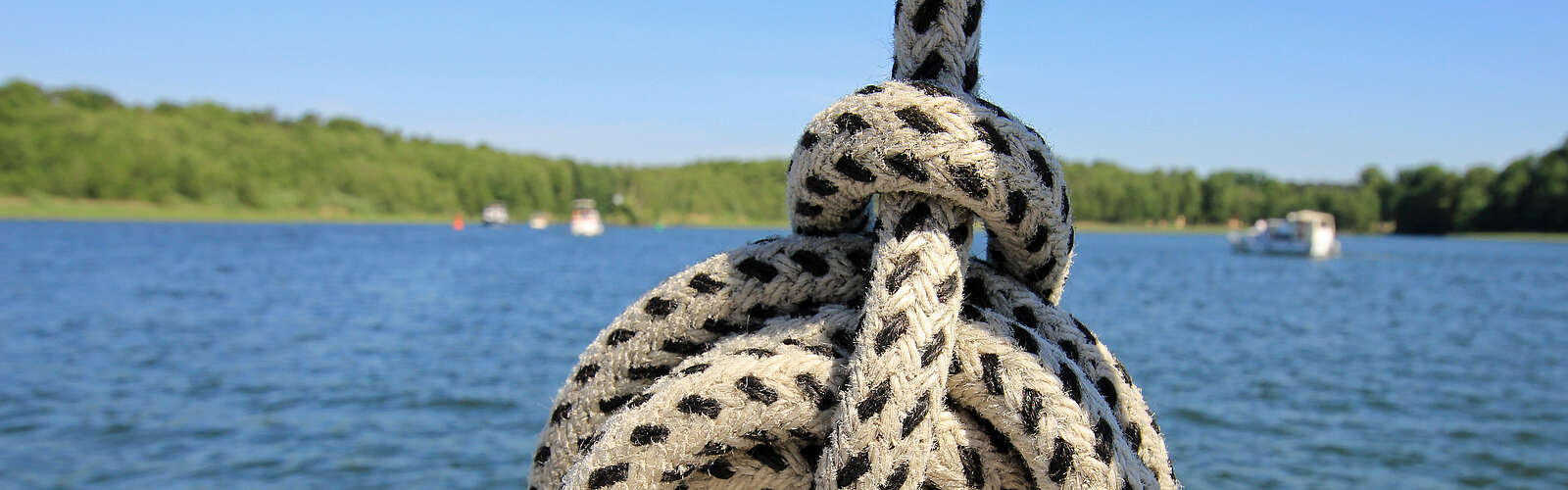 Ein Seemannsknoten in einem weißen Seil mit schwarzen Punkten. Im Hintergrund sieht man einen weiten See mit waldigem Ufer und mehrere Boote.