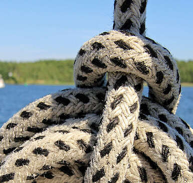 Ein Seemannsknoten in einem weißen Seil mit schwarzen Punkten. Im Hintergrund sieht man einen weiten See mit waldigem Ufer und mehrere Boote.